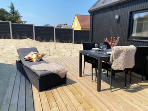 Um terraço com espreguiçadeira, mesa de jantar posta com pratos e copos, e um edifício de madeira escura ao fundo.