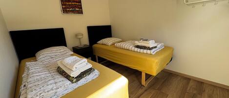A room with two beds, each with a yellow bedspread and white linens, a bedside table with a lamp, and a colorful abstract painting on the wall.
