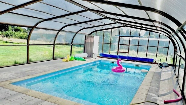An indoor swimming pool with a transparent roof, surrounded by a tiled floor and equipped with a pool ladder and a pink flamingo float. - Maison spacieuse avec piscine (Condé-sur-Risle)