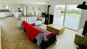 A spacious living area with a sofa, a red throw blanket, and a wooden coffee table. A kitchen with white cabinets and a dining area with chairs is visible in the background. - Maison spacieuse avec piscine (Condé-sur-Risle)