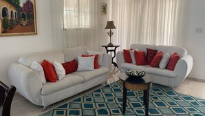 Un salon avec deux canapés blancs, des coussins rouges et blancs, un tapis  à motifs, une petite table et un tableau accroché au mur.