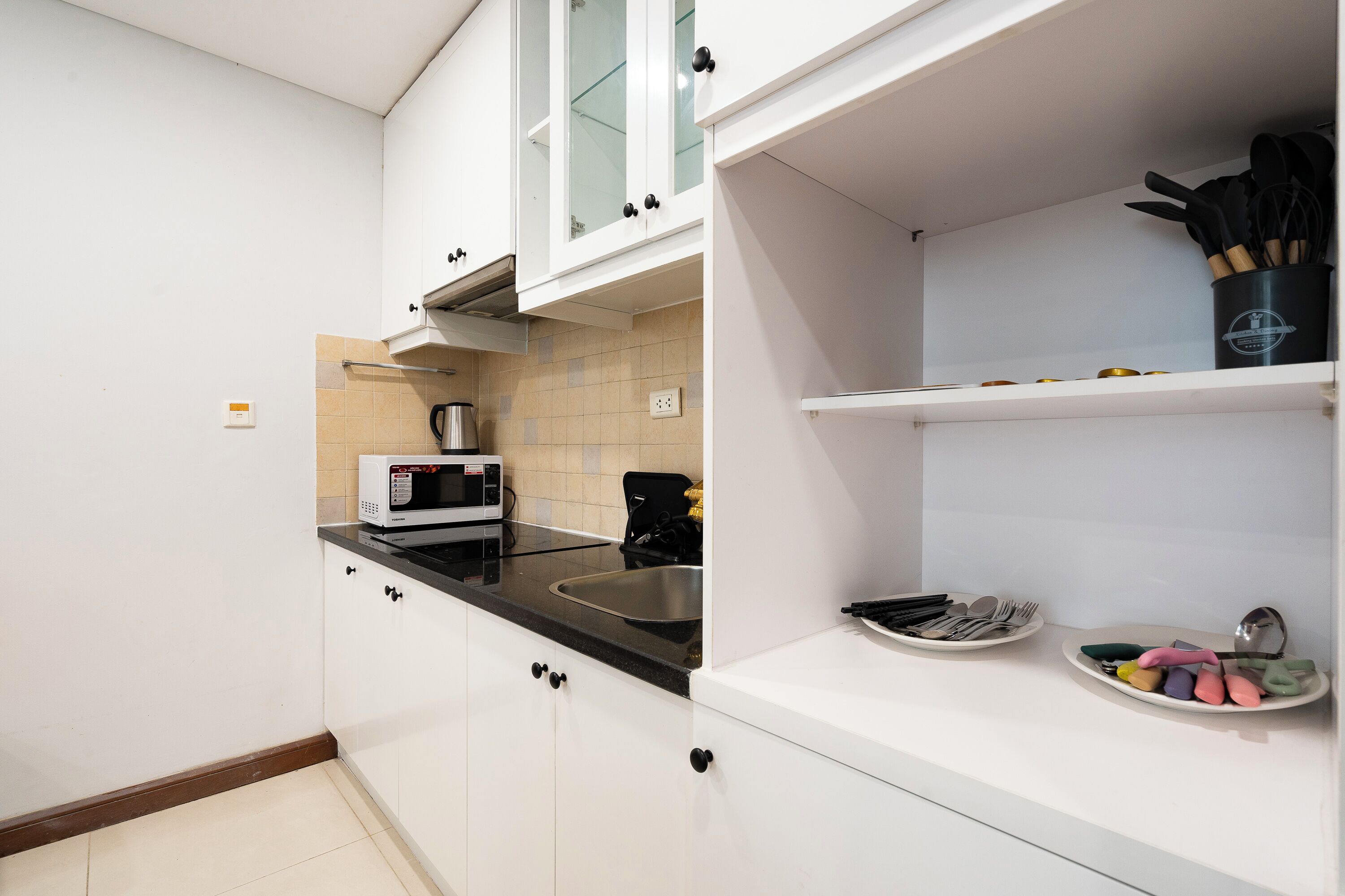 A modern kitchen with white cabinets, a black countertop, and a microwave. There are utensils on a shelf and a sink with a faucet.