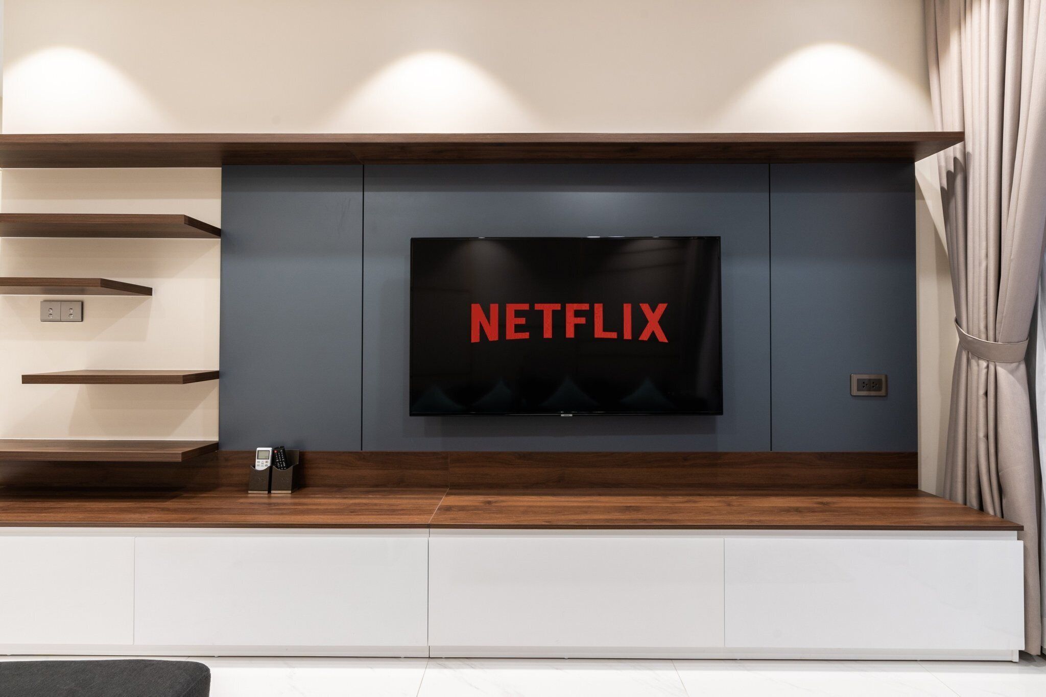 A modern living room with a flat-screen TV mounted on a grey wall, wooden shelves, and a built-in bench.