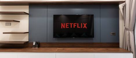 A modern living room with a flat-screen TV mounted on a grey wall, wooden shelves, and a built-in bench.