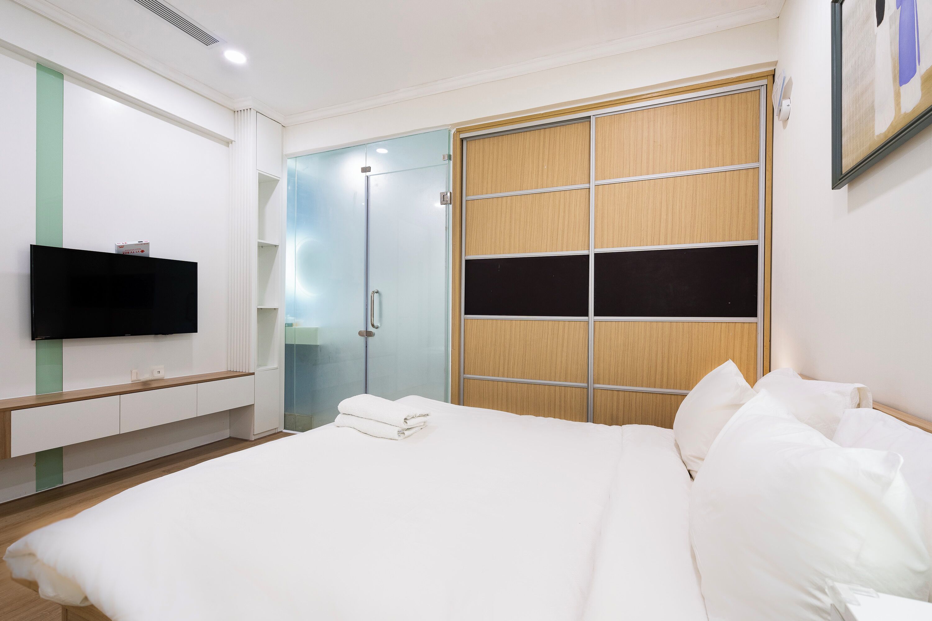 A modern hotel room with a large bed, a flat-screen TV mounted on the wall, a wooden wardrobe, and a glass shower enclosure.