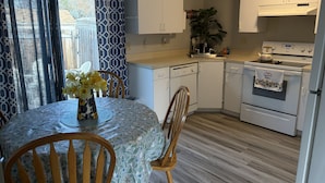 A compact kitchen with white cabinets, a dishwasher, an oven, and a sink. There's a dining table with chairs and a vase of flowers. - Charming, Cozy Boise Duplex (Boise)