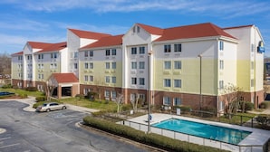 A multi-story hotel with a swimming pool, parking lot, and a clear blue sky.