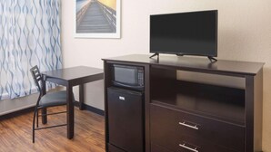A compact room with a desk, a chair, a microwave, a flat-screen TV, and a framed picture of a pier.