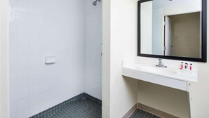 A bathroom with white tiled walls, a shower, a sink with a mirror, and a small shelf with toiletries.