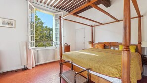 A four-poster bed with a wooden frame and a yellow patterned bedspread. There is a window with a view of greenery outside, and a radiator on the floor. - Centre Ville à Pied Dans une Authentique Bastide Xviieme Rénovée (Aix en Provence)