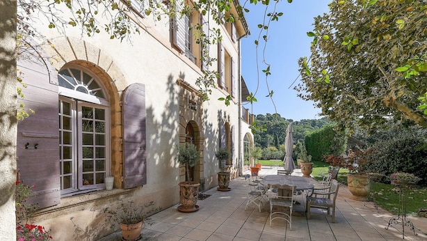 A courtyard with a tiled patio, outdoor furniture, potted plants, and a building with arched windows and shutters. - Centre Ville à Pied Dans une Authentique Bastide Xviieme Rénovée (Aix en Provence)
