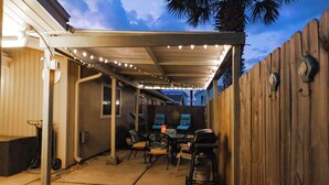 Ein überdachter Patio-Bereich mit Lichterketten, Gartenmöbeln und einem Grill.