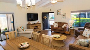 Ein geräumiges Wohnzimmer mit Holzesstisch, braunen Sofas, einem an der Wand montierten Fernseher und einem Deckenventilator.