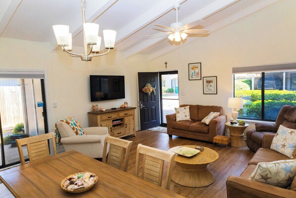A spacious living room with a wooden dining table, a brown sofa set, a television mounted on the wall, and a ceiling fan.