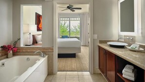 Un baño moderno con bañera, lavamanos y espejo. Se ve una cama a través de una puerta abierta que da a un balcón con vistas a zonas verdes.