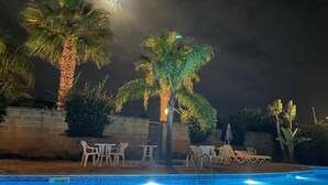 Uma área de piscina à noite, com palmeiras, espreguiçadeiras e piscina revestida de azulejos.