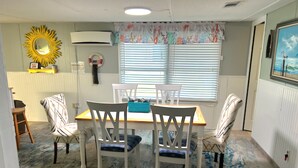 Een eethoek met een tafel en vier stoelen, een zonnespiegel, een airconditioning en een raam met jaloezieën.