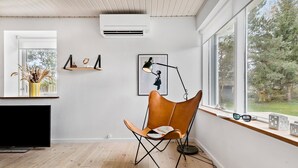 Una habitación moderna con suelo de madera, un ventanal con vistas a árboles, aire acondicionado integrado en la pared, un estante con objetos decorativos y una silla de diseño original.