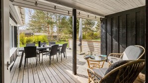 Un porche techado al aire libre con una mesa y sillas, una silla de mimbre, y vistas a un jardín y una casa al fondo.