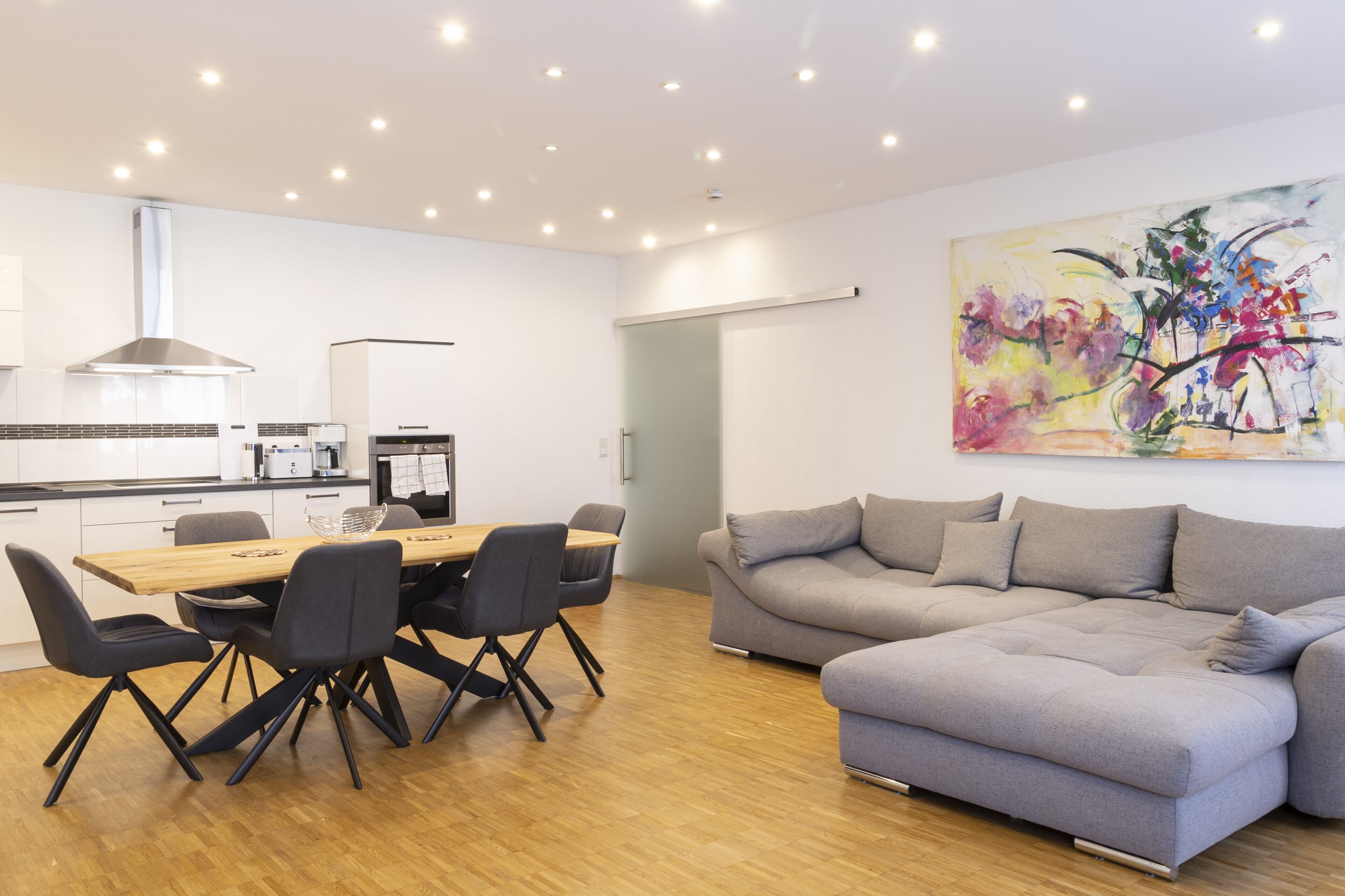 A modern kitchen with a dining area, a gray sofa, and a colorful abstract painting on the wall.