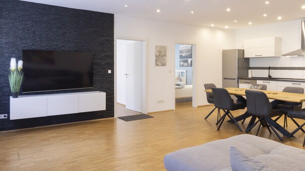 A modern living room with a flat-screen TV, a dining area with a wooden table and chairs, and a kitchen with white cabinets and stainless steel appliances. - Haus Oselbach- Gemütlich, Zentral, Modern (Zweibrücken)