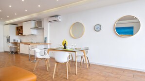Een moderne keuken met witte kastjes, een ingebouwde oven en een airconditioner. Er is een eethoek met een glazen tafel, witte stoelen en een geel decoratief object.