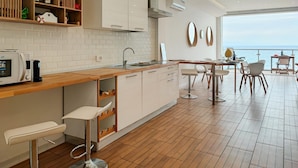 Een moderne keuken met witte kasten, een houten aanrecht en een barkruk. Er is een zithoek met een tafel en stoelen, en uitzicht op de zee door grote ramen.
