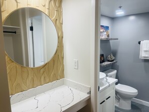 A bathroom with a marble countertop, a round mirror, a white toilet, and a shelf with decorative items. - Parking Included Upgraded w King Bed Marine Surf Waikiki (Honolulu)
