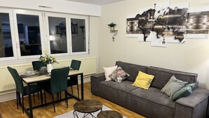 A modern living room with a dining area, featuring a grey sofa, green chairs, a wooden dining table, and a black and white striped rug. - Charmant Appartement à Metz - Proximité Gare (Metz)
