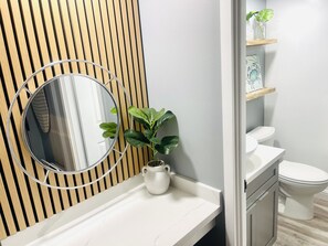 A bathroom with a wooden slat wall, a round mirror, a potted plant, a white sink, and a toilet.