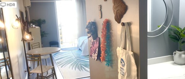 Ein Hotelzimmer mit einem Bett, einem runden Spiegel, einem kleinen Tisch und einem Stuhl. Auf dem Fensterbrett steht eine Pflanze und an der Wand hängt ein Bild mit dem Schriftzug "Hawai Ocean Club".