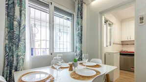 A dining area with a table set for four, two windows with curtains, a kitchen visible in the background, and a small plant on the table. - Nice apartment in Santa Pola with WiFi (Santa Pola)