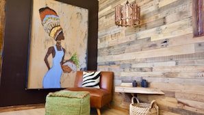Una habitación con pared de madera, un sofá de cuero marrón, un otomán tejido verde, un cojín estampado con motivos de cebra, una cesta tejida y un cuadro de una mujer con vestido azul.