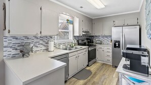 A modern kitchen with white cabinets, a stainless steel refrigerator, a coffee machine, and a window with a view of buildings outside. - Tranquil Breeze-5 min. walk to the beach! Book now for great fall/winter rates! (Oak Island)