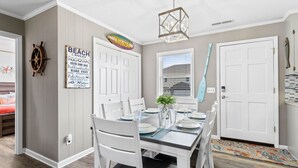 A dining area with a white table set for four, a potted plant, and a wall-mounted sign reading "BEACH RULES". - Tranquil Breeze-5 min. walk to the beach! Book now for great fall/winter rates! (Oak Island)