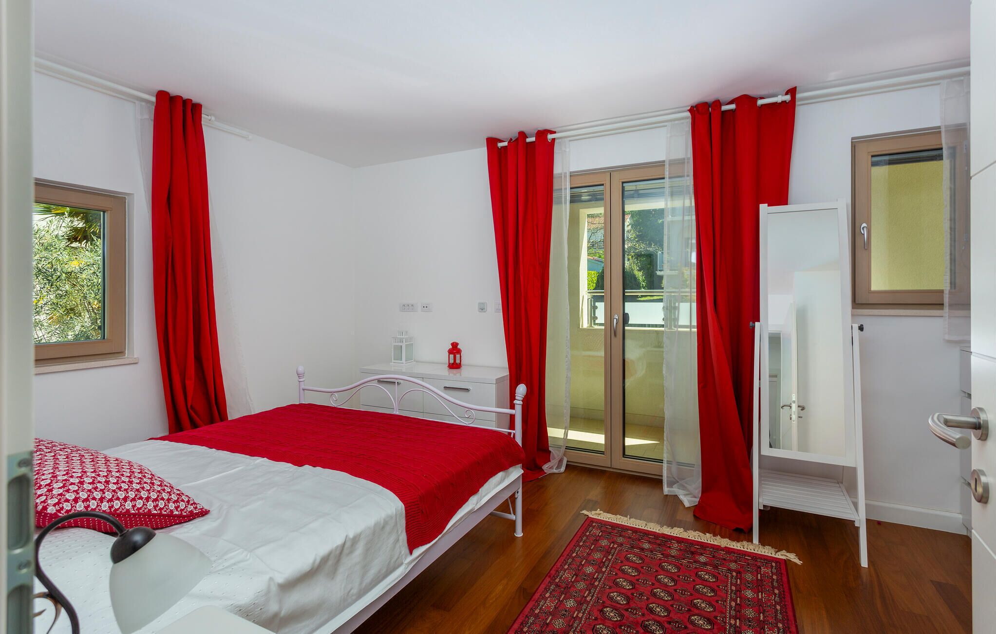 A bedroom with a bed, a bedside table, a lamp, a red rug, and a sliding glass door leading to an outdoor area.
