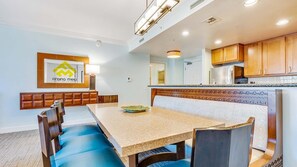 A dining area with a table, chairs, and a bench. There is a kitchen area with wooden cabinets and a refrigerator. - 𝐅𝐢𝐧𝐝𝐢𝐧𝐠 𝐌𝐞𝐢𝐥𝐢’𝐬 Beach Fun Waiohai Club Resort + Amenities (Koloa)