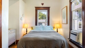 A bedroom with a bed, two bedside lamps, a dresser, a window with an air conditioner, and a wall-mounted heart decoration. - Cozy and charming. Your home away from home in the heart of Viroqua. (Viroqua)