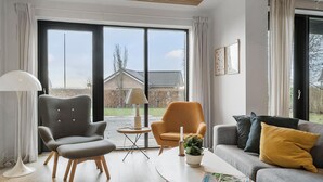 A modern living room with a grey armchair, an orange armchair, a grey sofa with a yellow pillow, and a white coffee table. There is a floor lamp, a small table with a candle and a plant, and a window with curtains. - Awesome home in Haderslev with WiFi (Haderslev)