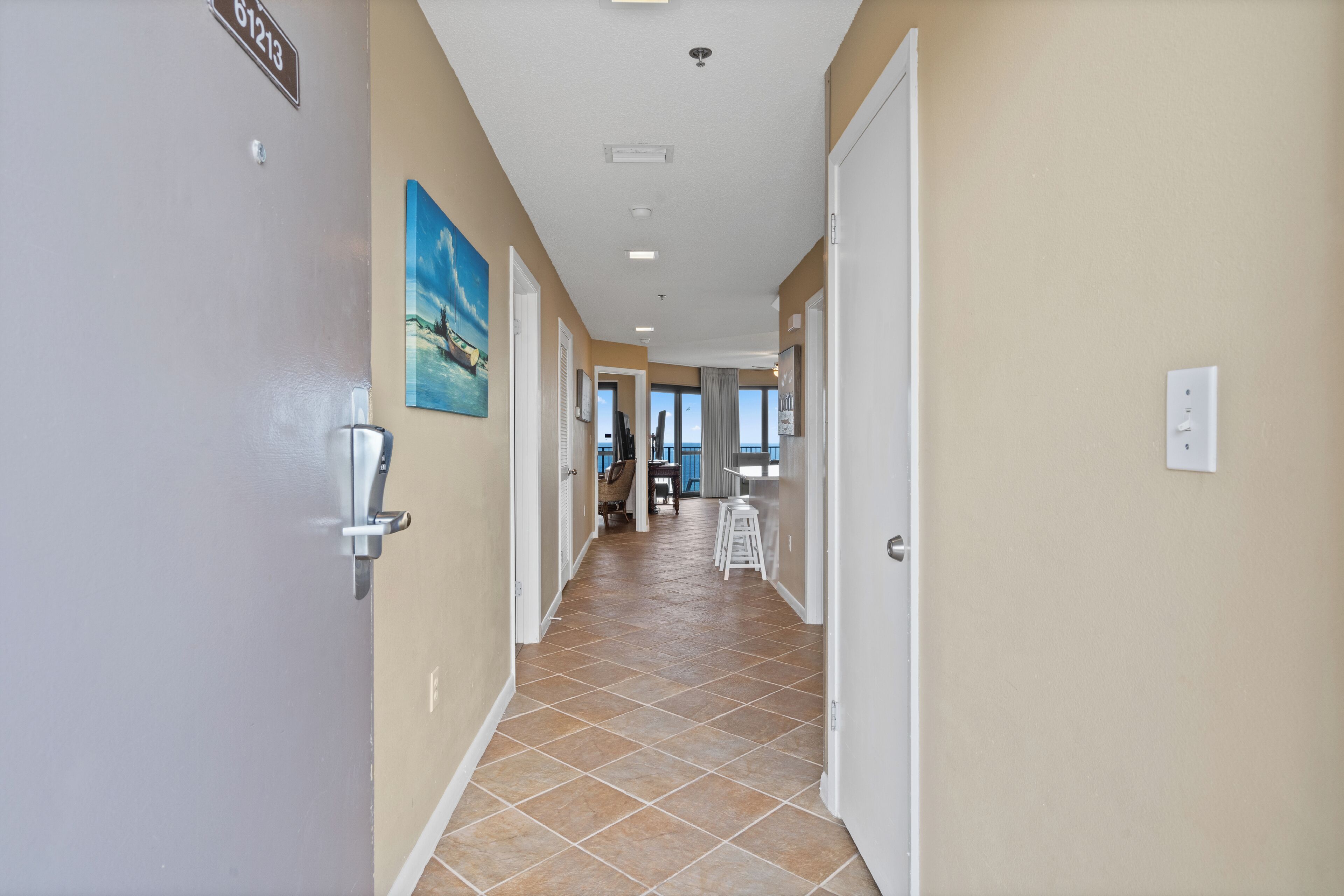 A hotel hallway with beige walls, a tiled floor, and a view of the ocean through large windows.
