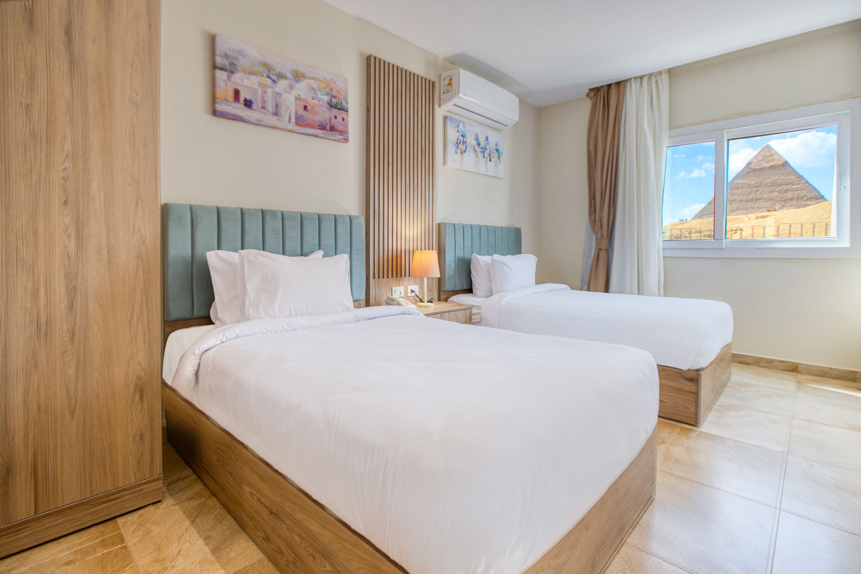 A hotel room with two beds, a wooden wardrobe, a nightstand, and a window with a view of a pyramid.