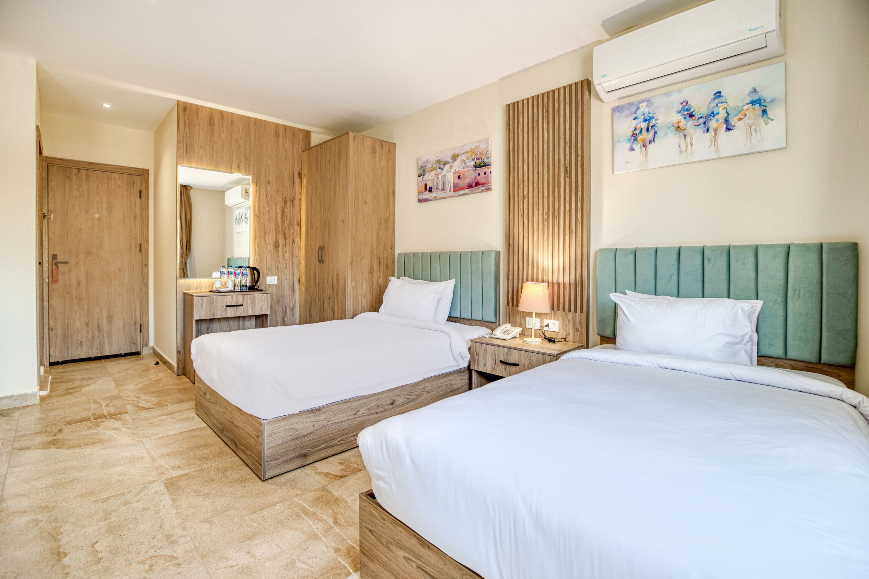 Habitación de hotel con dos camas, cabeceros de madera, una mesita de noche con lámpara, un espejo y obras de arte en la pared.