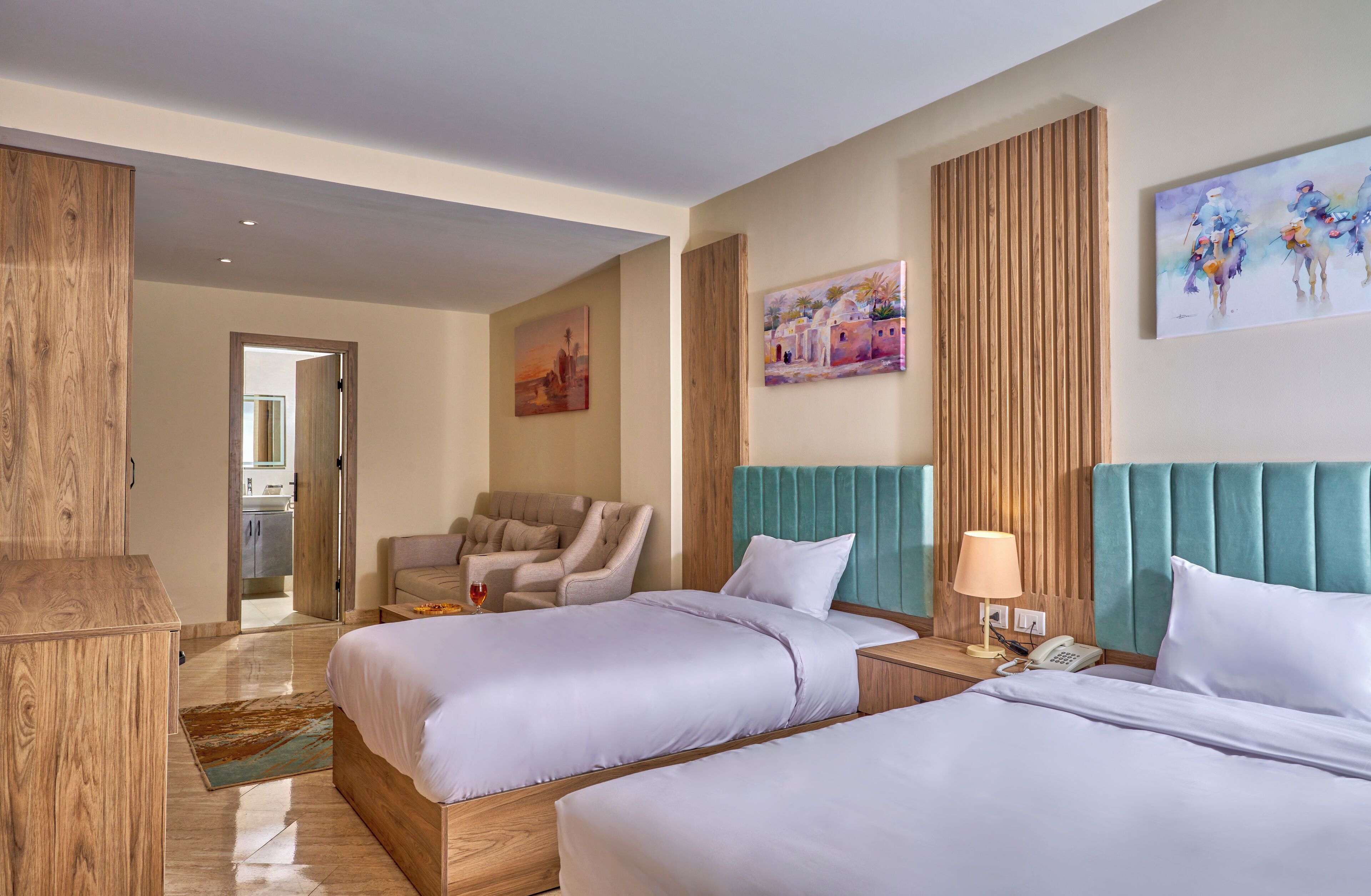 A hotel room with two beds, a wooden headboard, a sitting area with a chair, a painting on the wall, and a wooden panel wall.