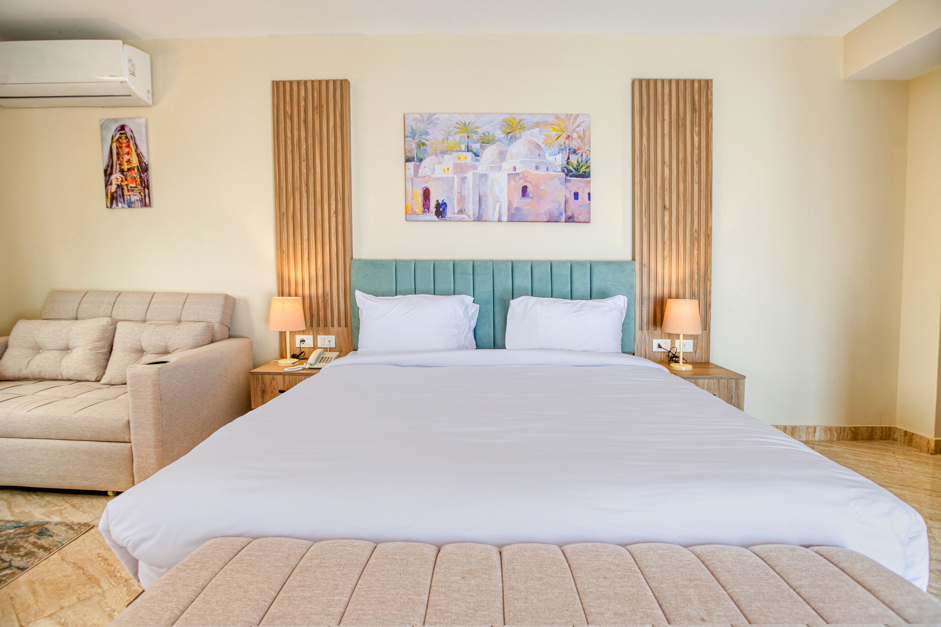 A hotel room with a large bed, a sofa, two nightstands, a wall-mounted air conditioner, and a painting of a village scene.