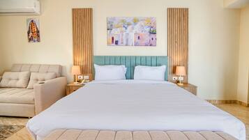 A hotel room with a large bed, a sofa, two nightstands, a wall-mounted air conditioner, and a painting of a village scene.