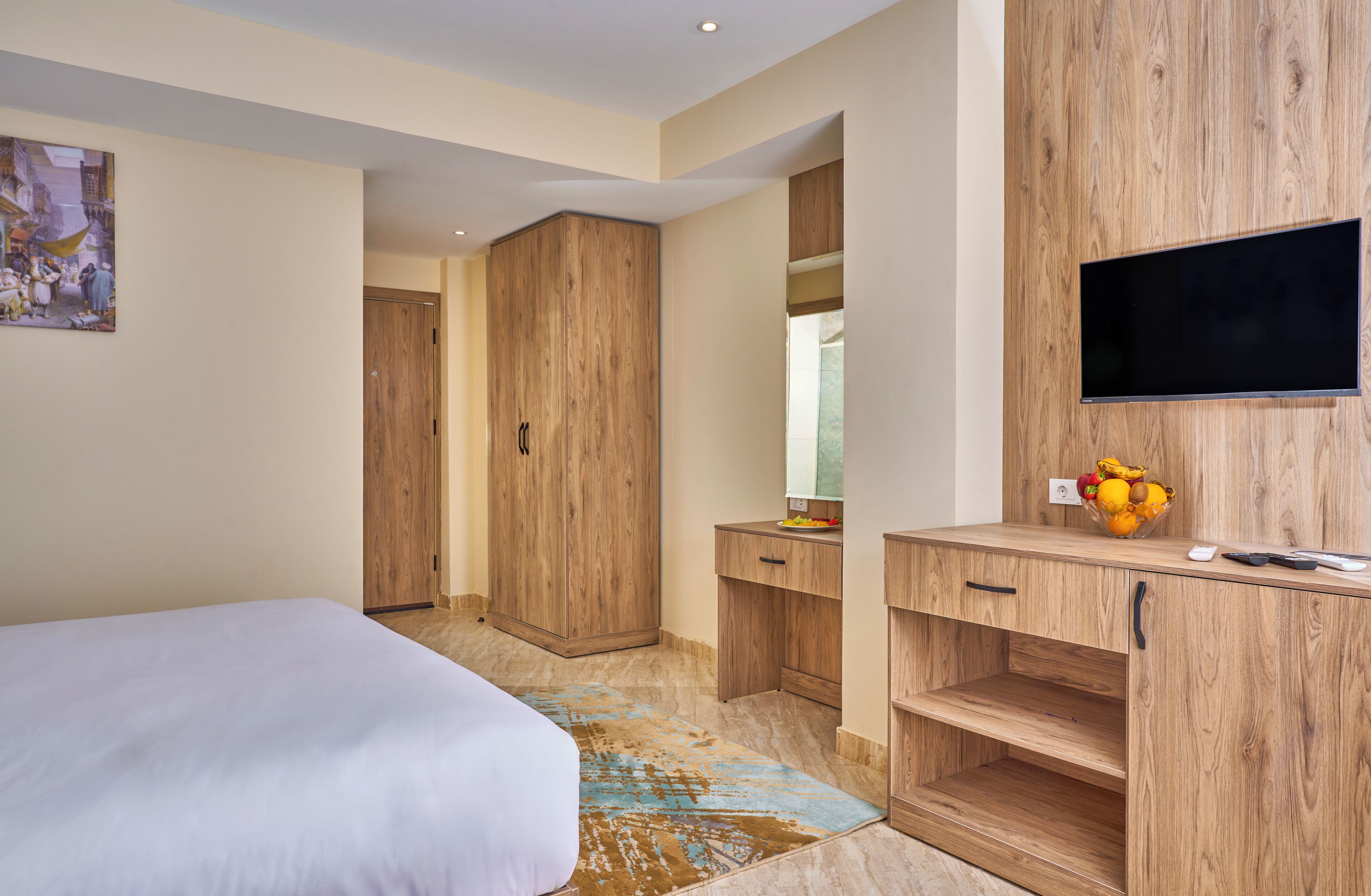 A modern bedroom with a large bed, wooden wardrobe, a desk with a flat-screen TV, and a fruit bowl on the desk.