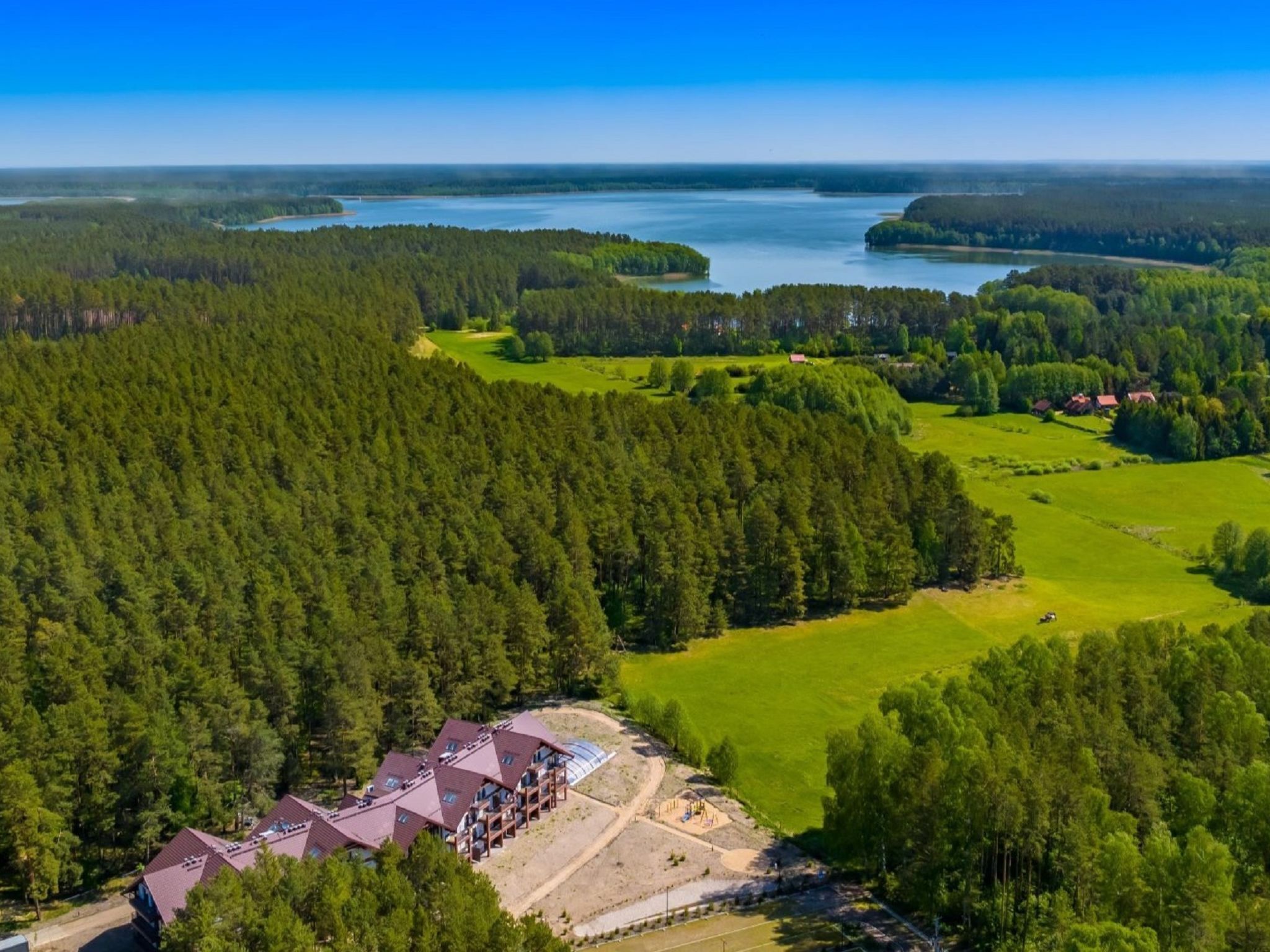 Vista aérea de una zona boscosa con un edificio que cuenta con varias construcciones de techo rojo, rodeado de campos verdes y un lago al fondo.