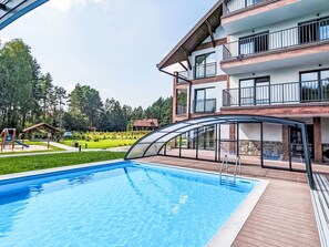 Une piscine entourée d’une enceinte en verre, bordée d’une terrasse en bois, et un immeuble à plusieurs étages avec des balcons en arrière-plan.