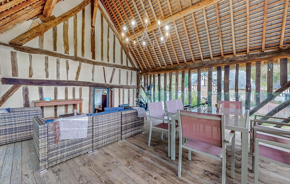 A rustic interior with exposed wooden beams, a high ceiling, wicker furniture, and large windows offering a view of the outside.