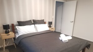 A neatly made bed with a dark bedspread and white pillows, a wooden nightstand with a lamp, and a door to the right.